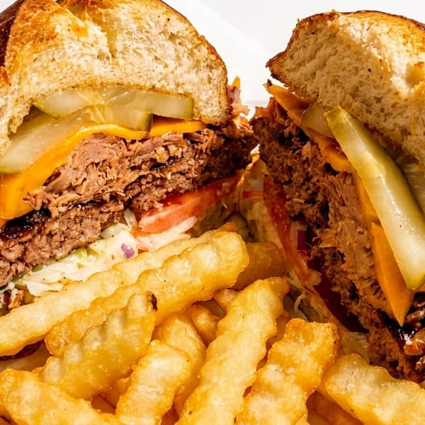 A large burger cut in half, with cheese, pickles, and pulled pork, served with crinkle-cut fries.